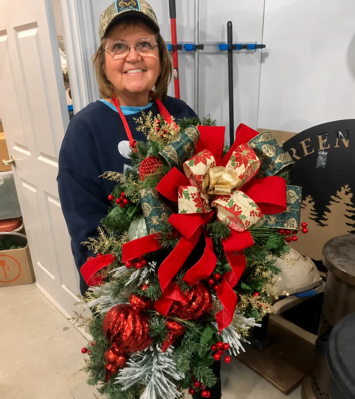 Smiling woman holding a large handmade Christmas wreath and greenery arrangement with red ribbon.
